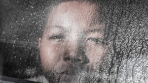 A sad young boy gazing out a rain streaked window