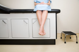 Female patient sitting on doctor's table in gown, with ankles crossed.