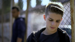 Upset teen boys leaning on metal fence, abandoned by society.