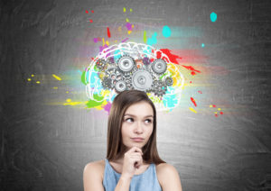 Portrait of a dreamy teen girl wearing a blue dress and holding her hands near the face. She is standing near a blackboard with a colorful brain sketch and gears on top of it