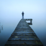 Depressed man standing alone on a jetty on a foggy autumn day.