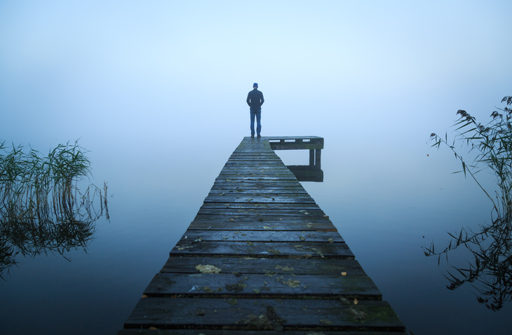 Depressed man standing alone on a jetty on a foggy autumn day.